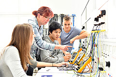 Eine Gruppe junger Menschen sitzt vor einer Wand, an der technische Geräte hängen. Eine Frau steht hinter ihnen.