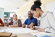 Drei Personen mit Migrationshintegrund sitzen gemeinsam an einem Tisch. Eine Person schreibt auf einen großen Block.