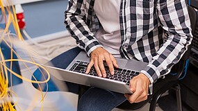 Person sitzt auf einem Stuhl und hat einen Laptop auf dem Schoß, im Vordergrund sind Kabel zu sehen.