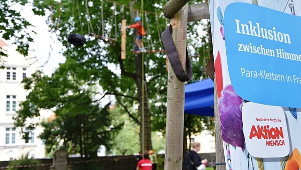 Inklusives Para-Kletterangebot: Im Kletterpark unterstützt eine Person eine kletternde Person, ein Schild der Aktion Mensch ist rechts im Bild zu sehen.