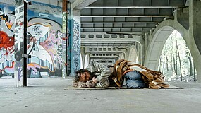 Ein Mann liegt unter einer Brücke auf dem Boden und schläft. Er ist mit einer Jeans und einer grauen Jacke bekleidet und teilweise von einer Decke bedeckt. Die Wände im Hintergrund sind teilweise mit Graffitis besprüht. 