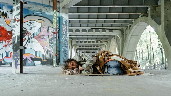 Ein Mann liegt unter einer Brücke auf dem Boden und schläft. Er ist mit einer Jeans und einer grauen Jacke bekleidet und teilweise von einer Decke bedeckt. Die Wände im Hintergrund sind teilweise mit Graffitis besprüht. 