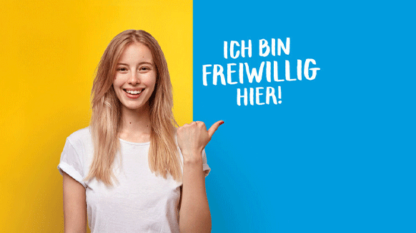 Junge Person mit weiblichem Erscheinungsbild vor einem gelb-blauen Hintergrund mit der Aufschrift "Ich bin freiwillig hier".