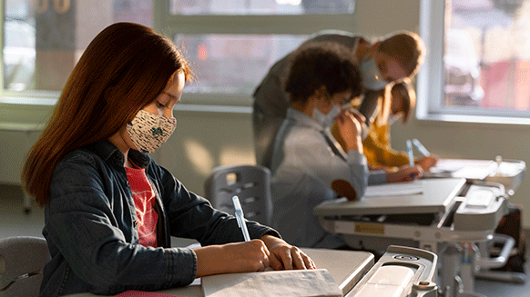 Mehrere Schüler*innen, die Masken tragen, sitzen in einem Klassenzimmer und schreiben in Schulhefte.